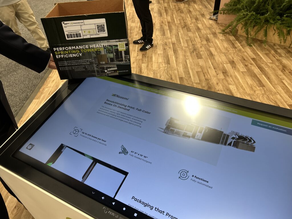 Packsize revealed its EFI X5 Nozomi system at Pack Expo Las Vegas, capable of producing full-color boxes on demand. EFI representatives told Packaging OEM that the system replaces the need for label barcodes, photographed here next to a digital image of the machine.