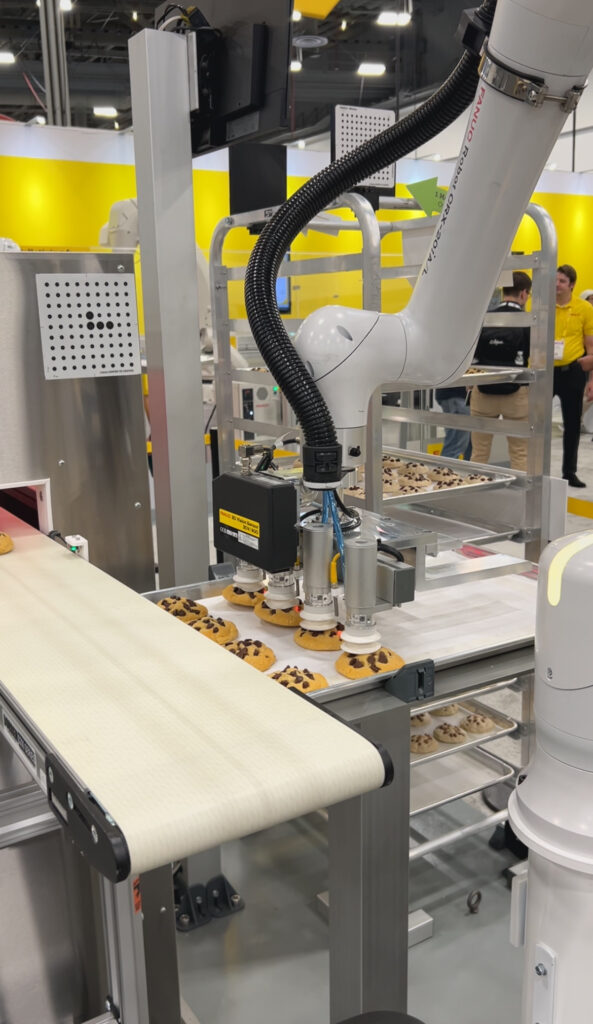 Fanuc highlighted CRX food-grade cobots at Pack Expo 2025 at the Las Vegas Convention Center. Dual CRX-20iA/L robots load chocolate chip cookies onto a conveyor, place them on trays, and move trays onto carts using Fanuc iRVision One Marker Offset. The cobots use NSF-H1 food-safe grease, white epoxy coating, and are IP67 rated for hygienic handling and reduced manual labor in bakery operations.