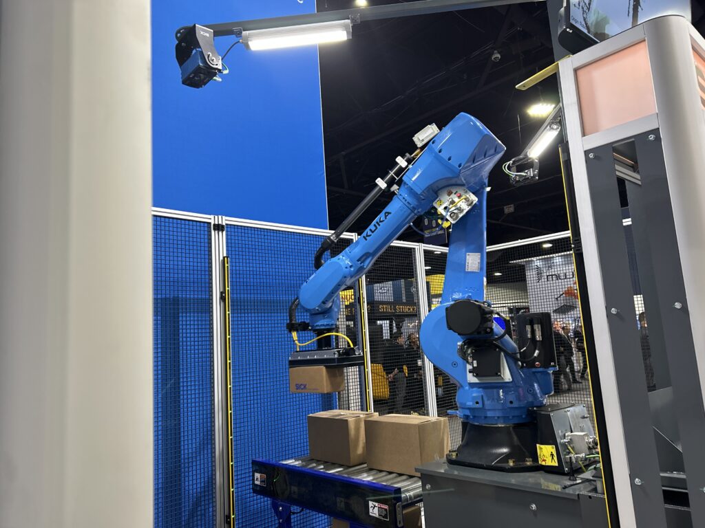 A KUKA robot palletizing boxes at the SICK booth at Modex 2026 in Atlanta, Georgia, demonstrating AI-enabled automation in a physical handling application.
