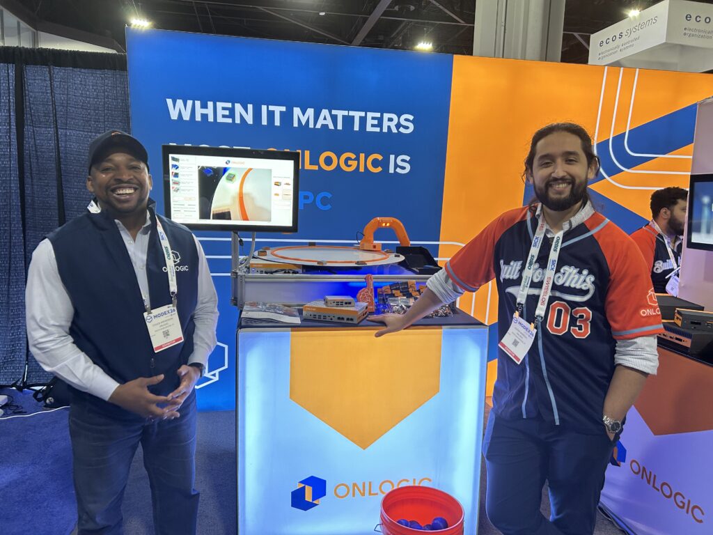 Chuck Nwokocha, sales director at OnLogic, and Alexis Mangual, business development manager at OnLogic, at Modex 2026 in Atlanta, Georgia, standing next to a machine vision demo that sorted items by color.